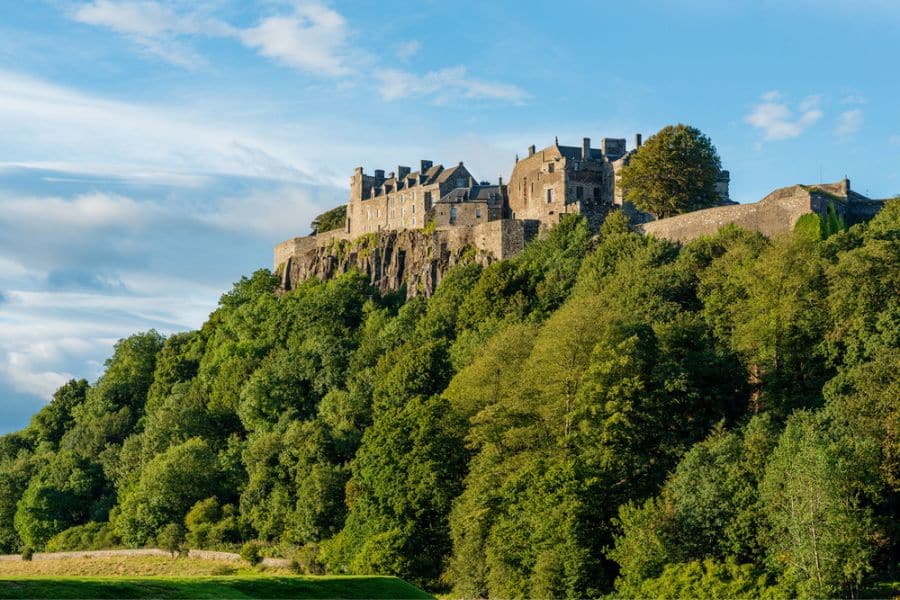 Stirling Castle sits high on a rocky hilltop surrounded by dense greenery, with golden sunlight illuminating its historic stone walls.