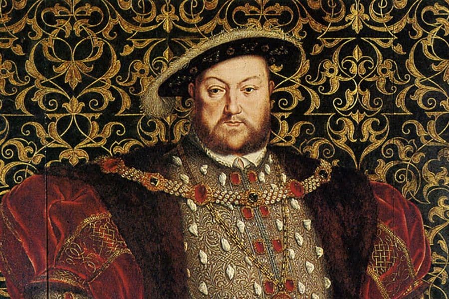Portrait of Henry VIII wearing an opulent fur-trimmed robe adorned with large jewels and a feathered hat, posed against an ornate gold and black tapestry. This image represents the controversial Tudor king who faced multiple assassination plots during his reign.