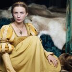 Actress in historical costume portraying Anne Neville in a gold gown, seated on a fur-covered bed with a solemn expression, from a modern TV adaptation.