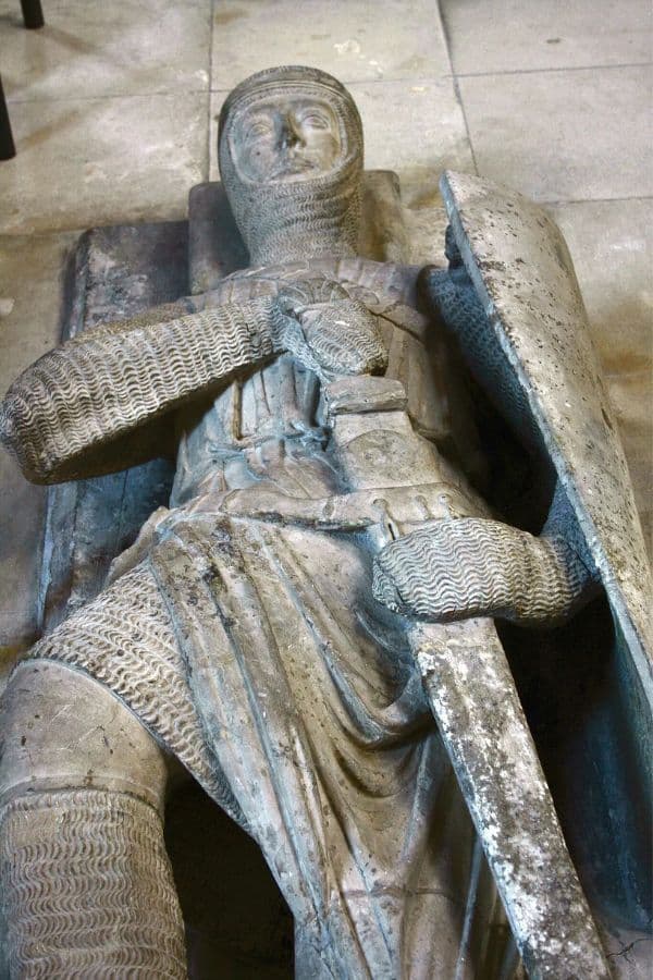 Close-up of a medieval stone effigy of a knight in full chainmail armor, hands on a sword resting on his chest, with a large shield on his left side. The detailed sculpture lies on a tomb inside a stone chapel or church.