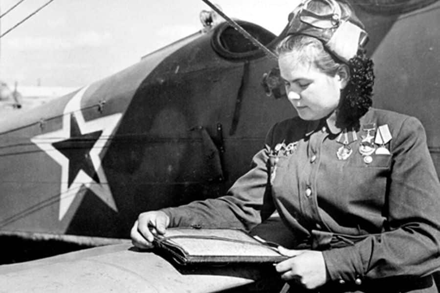 A decorated Soviet woman pilot, part of the Night Witches, reads from a book while resting on the wing of a biplane marked with a red star. She wears a pilot’s helmet and several medals on her jacket.