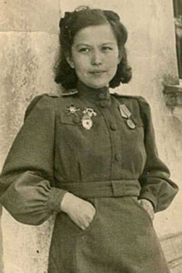 A young Soviet woman in uniform, identified as a Night Witch pilot, leans against a building wall with hands in her pockets. She wears military medals on her chest and has a calm, confident expression.