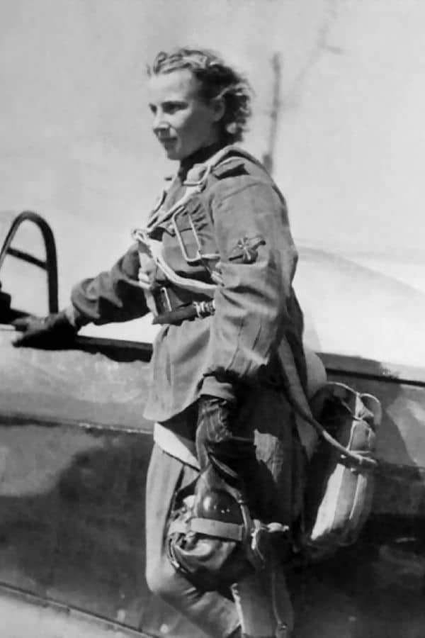 A female Soviet aviator from the Night Witches stands beside a military aircraft, dressed in a flight suit with gloves and a parachute harness. She looks into the distance, appearing focused and ready for flight.
