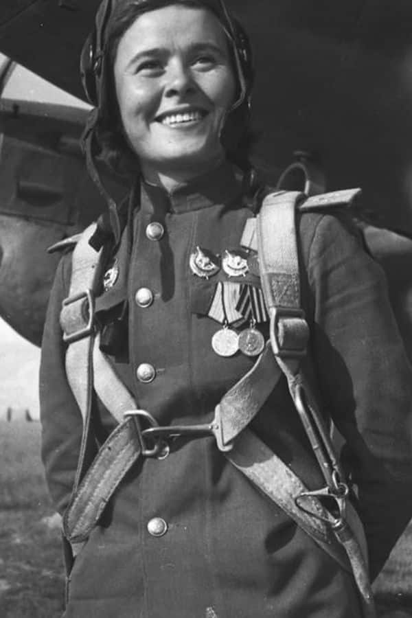 A smiling female Soviet pilot in a decorated uniform and parachute harness stands in front of her aircraft. As one of the Night Witches, she flew nocturnal bombing raids against the Nazis.