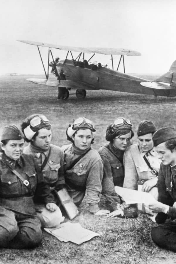 A group of Soviet women pilots from the Night Witches sit on the ground in front of a biplane, reviewing mission plans or flight documents. They wear flight uniforms and headgear, focused on their briefing.