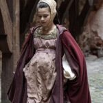 Young woman in a Tudor-style gown and headdress, visibly pregnant and walking across a medieval courtyard, representing a dramatized depiction of Margaret Beaufort during her early marriage to the first of her four husbands.
