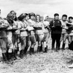 A row of Soviet women aviators from the Night Witches stand in a semicircle on open ground, holding notebooks and listening to a woman officer. Most wear light-colored uniforms and some have flight helmets or parachutes.