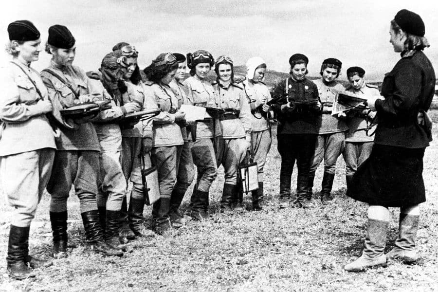A row of Soviet women aviators from the Night Witches stand in a semicircle on open ground, holding notebooks and listening to a woman officer. Most wear light-colored uniforms and some have flight helmets or parachutes.