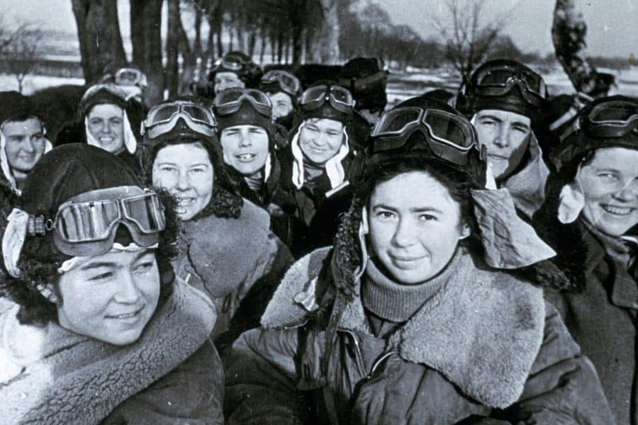 A group of young Soviet women pilots smile in cold weather gear and flight goggles, gathered outdoors during winter. These women were part of the legendary Night Witches, an all-female bomber regiment in World War II.