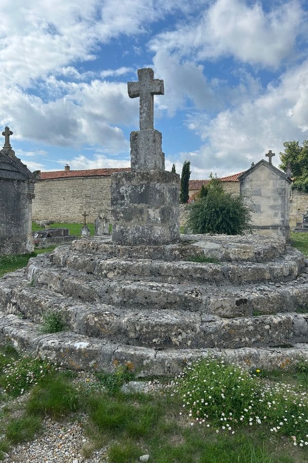 A weathered stone cross on a circular, tiered base stands in the middle of an old cemetery with medieval architecture in the background. The structure may be a Templar memorial or grave marker.