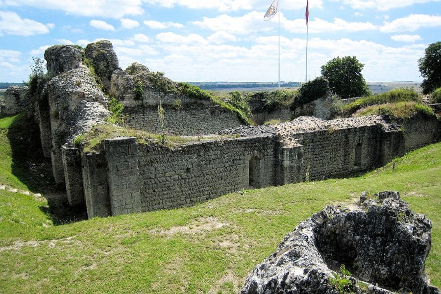 Ruins of a medieval stone castle at Fontevraud-l'Abbaye under a sunny sky, with two flags flying in the background. This historic site is linked to Juliane Fontevrauld, whose life intersected with the power struggles of Norman England.