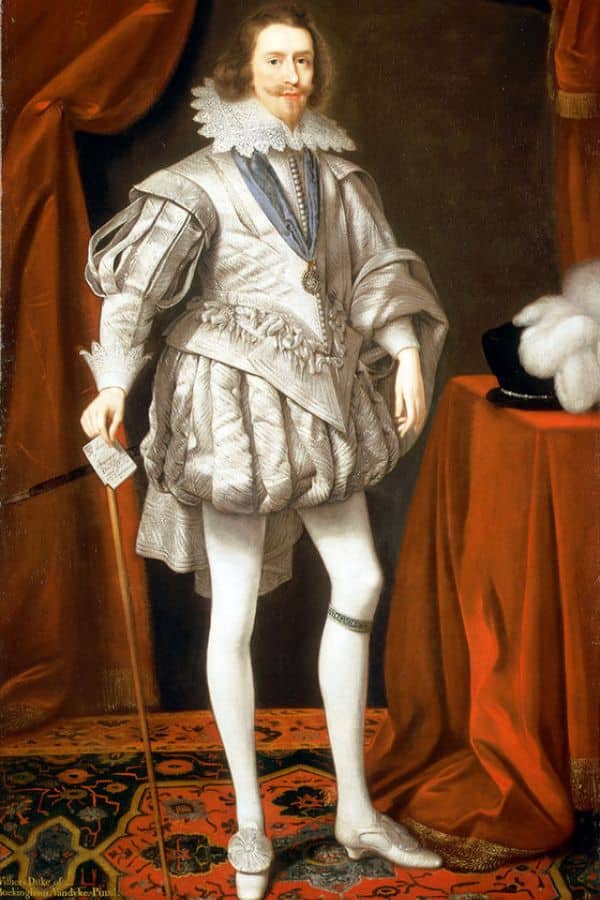 Full-body portrait of George Villiers dressed in an elaborate silver doublet and hose, holding a cane and standing on a richly patterned carpet. His white satin outfit and posture underscore his wealth and courtly prominence during the reign of King James I.