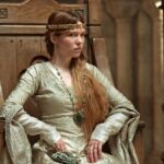 A woman dressed as Isabella of Angouleme sits sternly on a medieval throne, adorned with a gold circlet, long flowing hair, and an ornate gown with green gemstones. This cinematic representation captures her elegance and political strength as queen consort.
