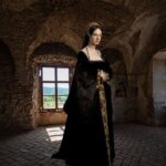 Woman dressed in an elaborate black and gold Tudor gown with a fur-lined cloak, standing in a dimly lit stone chamber with arched windows. This modern portrayal of Jane Boleyn evokes the atmosphere of Tudor England with period-accurate fashion and setting.