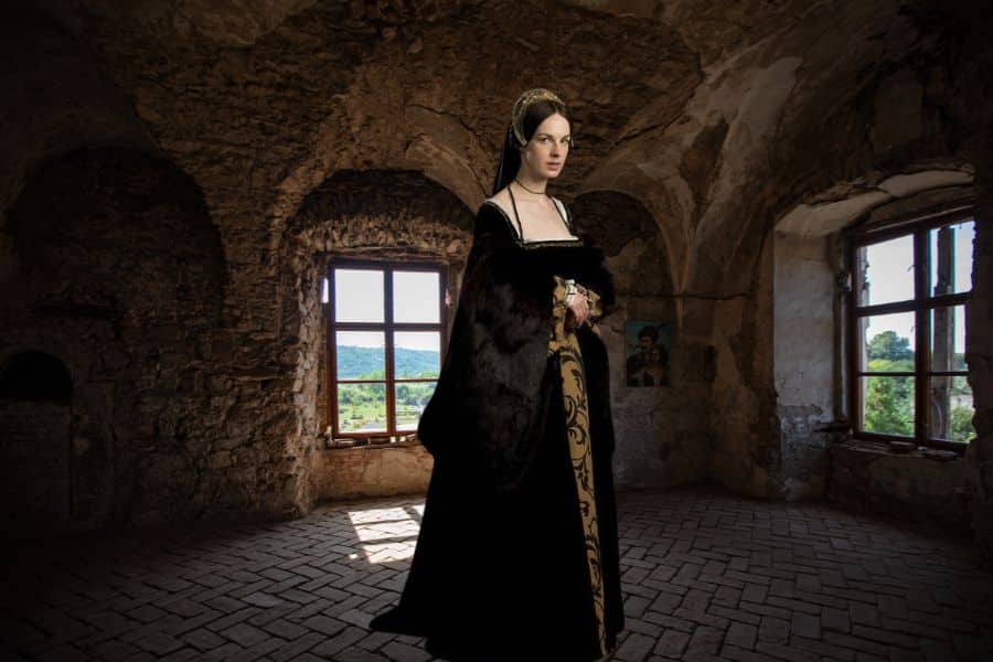 Woman dressed in an elaborate black and gold Tudor gown with a fur-lined cloak, standing in a dimly lit stone chamber with arched windows. This modern portrayal of Jane Boleyn evokes the atmosphere of Tudor England with period-accurate fashion and setting.