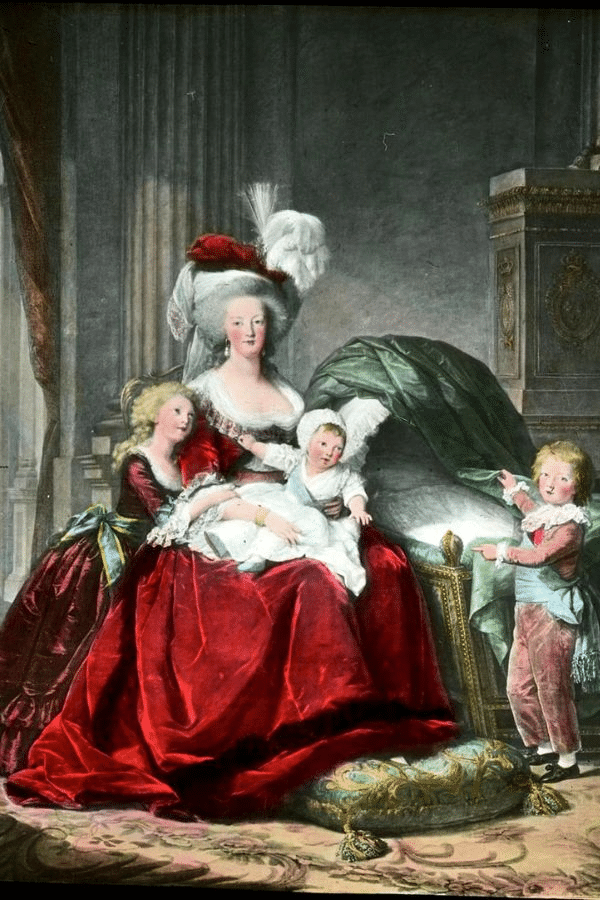 A regal portrait of Marie Antoinette seated with her children in a lavish palace interior, dressed in a red gown and white feathered hat. This image contrasts with the tragic tone of the last days of Marie Antoinette, highlighting her former royal status.