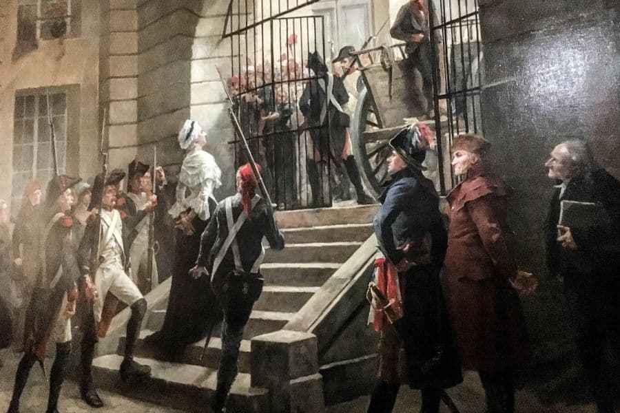 Marie Antoinette is shown under heavy guard entering the Conciergerie prison during the French Revolution, as crowds and soldiers surround her. This historic moment marks the beginning of the last days of Marie Antoinette.