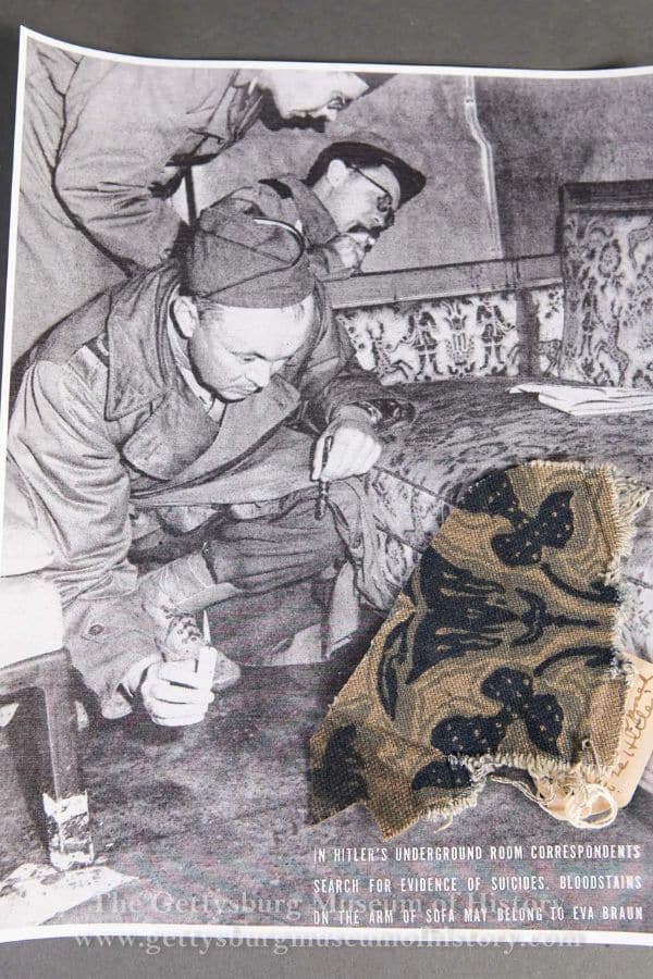 Black-and-white photo of Allied soldiers inside Hitler’s underground bunker in Berlin, closely examining a bloodstained couch. A piece of floral-patterned upholstery, possibly linked to Eva Braun, is pinned over the image. The printed caption notes a search for evidence of suicides. It's part of the new documentary regarding Hitler's DNA.