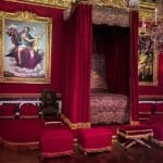A richly decorated bedroom in deep red and gold tones with a grand canopy bed behind velvet drapes, flanked by regal paintings and a crystal chandelier. This space showcases the extravagant design often found in royal palaces where separate bedrooms served ceremonial and private functions.