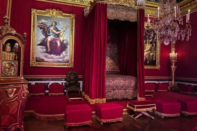 A richly decorated bedroom in deep red and gold tones with a grand canopy bed behind velvet drapes, flanked by regal paintings and a crystal chandelier. This space showcases the extravagant design often found in royal palaces where separate bedrooms served ceremonial and private functions.