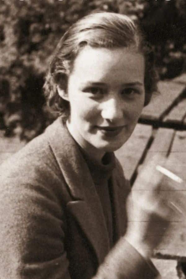 Vintage photo of a young Marie-Madeleine Fourcade sitting outdoors with a slight smile, holding a cigarette, capturing her early life before leading a major WWII resistance network.