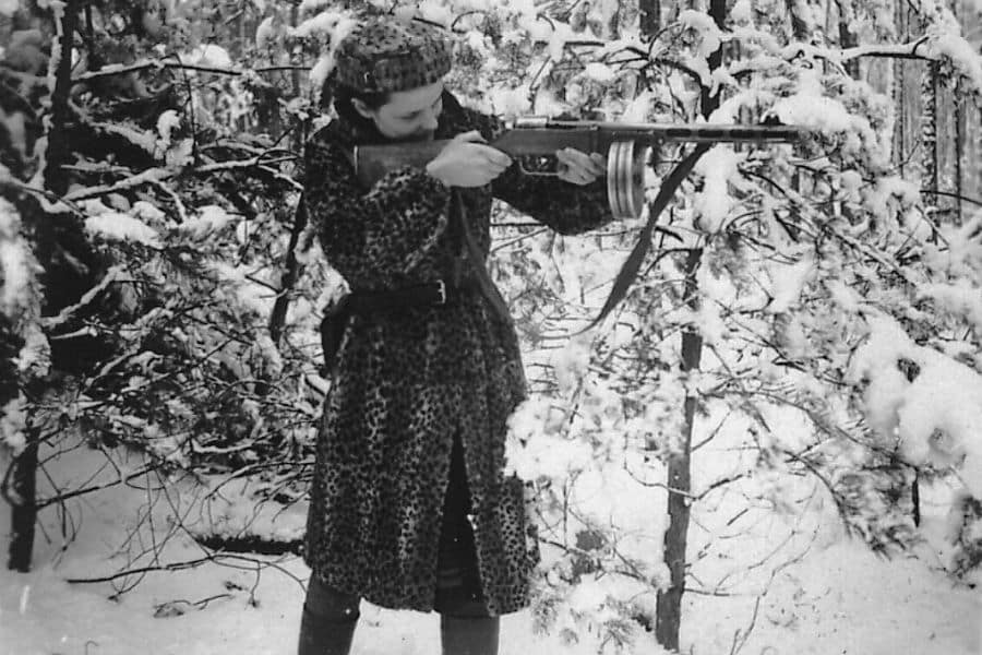 Faye Schulman, a Jewish partisan photographer and resistance fighter during World War II, stands in a snowy forest aiming a rifle while wearing a long coat and boots—an iconic image of her defiance and survival.