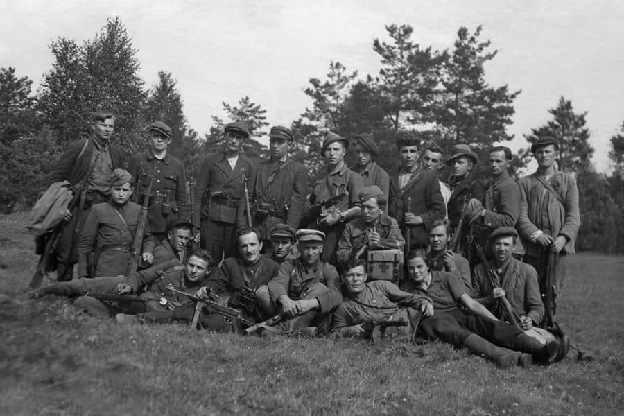A group of World War II Jewish partisans pose with rifles and other weapons in a forest clearing, showcasing the resilience and resistance of those who fought against Nazi forces during the Holocaust, including figures like Faye Schulman who documented such moments through photography.