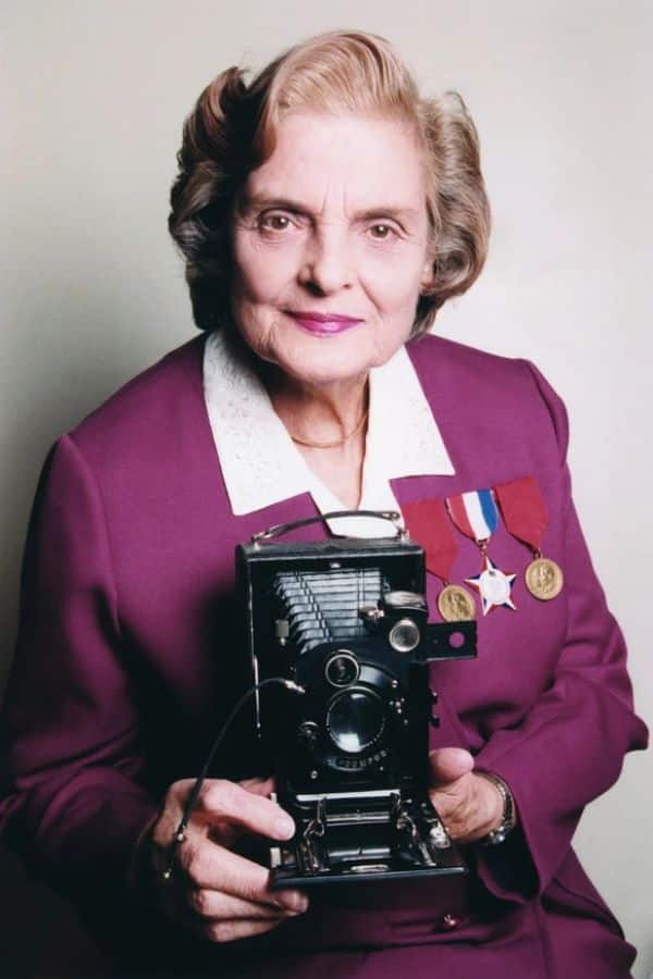 Faye Schulman, elderly and dignified, poses in a purple outfit adorned with medals, holding the vintage camera she used during World War II. A Jewish partisan and wartime photographer, she is celebrated for documenting resistance and survival during the Holocaust.