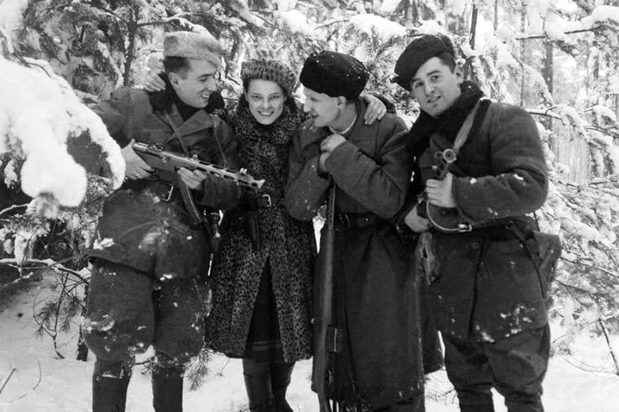 Faye Schulman, dressed in her signature leopard-print coat, stands smiling among three fellow Jewish partisans in a snowy forest during World War II. The group, armed and huddled together, displays rare warmth and camaraderie amid the harsh realities of resistance life.