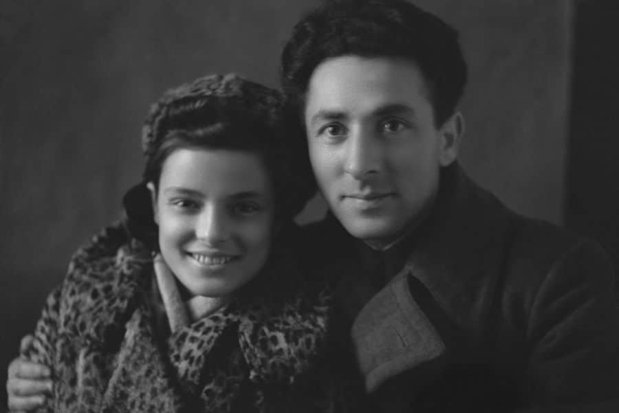 Faye Schulman, wearing her iconic leopard-print coat, smiles warmly alongside her husband Morris Schulman in a posed portrait. Both former Jewish partisans, they appear close and content, capturing a rare moment of peace and intimacy after surviving the horrors of World War II.