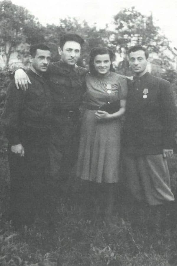 Faye Schulman stands proudly with her two brothers and husband after World War II, wearing a civilian dress and a medal pinned to her chest. The group poses closely in a grassy outdoor setting, symbolizing survival, resilience, and recognition of their wartime resistance efforts.