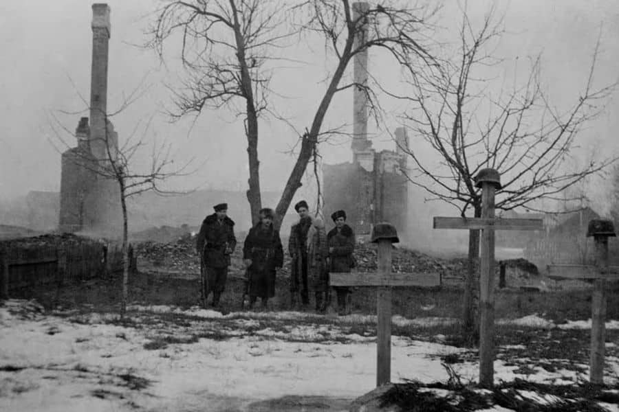 A somber scene of four Jewish partisans, including Faye Schulman, standing in a snow-covered graveyard with wooden crosses marked by military helmets. Behind them are the ruins of buildings and tall chimneys, evoking the destruction of war and the memory of those lost during the Holocaust.