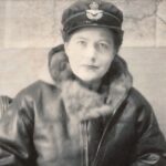 Vera Atkins in uniform wearing a fur-collared leather coat and officer's cap, seated against a wall map, reflecting her wartime intelligence work.