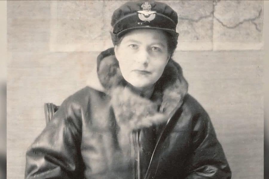 Vera Atkins in uniform wearing a fur-collared leather coat and officer's cap, seated against a wall map, reflecting her wartime intelligence work.