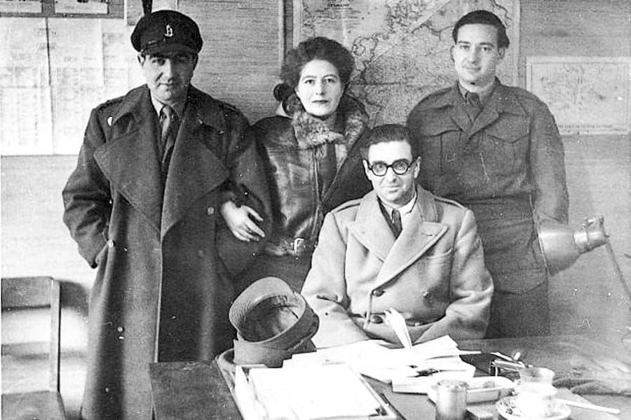 Historic black-and-white photo of Vera Atkins standing among three men in a WWII operations room with maps on the wall, showing her leadership in SOE missions.