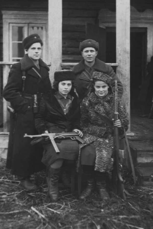 Faye Schulman (seated right), wearing a leopard-print coat and holding a rifle, poses with fellow Jewish partisans during World War II in front of a wooden building. This rare photograph captures the resilience and unity of the resistance fighters who opposed Nazi forces.
