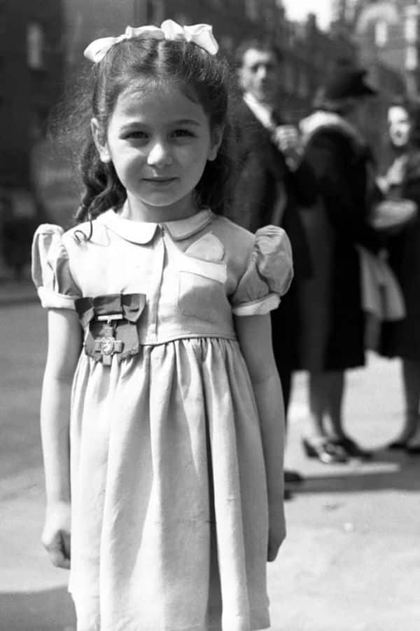 A young girl in a puff-sleeve dress with a ribbon in her hair stands in the foreground of a black-and-white street scene. This childhood photo captures the early life of Violette Szabo.