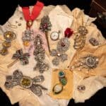 A collection of Empress Sisi's ornate jewelry displayed on cream fabric, including brooches, medallions, jeweled hairpins, and a pocket watch. Many pieces feature intricate gold, diamond, ruby, and emerald designs.