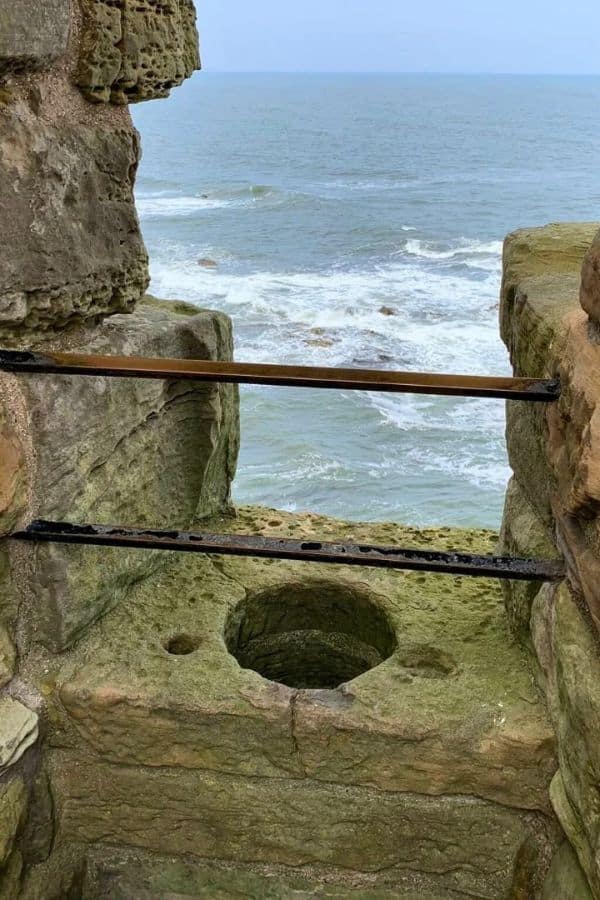 Stone toilet opening carved into a cliffside ruin with metal bars across the gap and rough ocean waves visible below. This photo shows a dramatic coastal example of medieval toilets built directly over the sea for waste drainage.