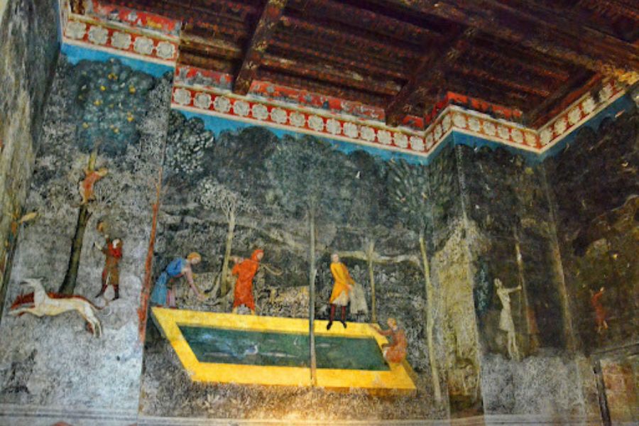 Ancient wall fresco painted inside a stone chamber depicting medieval figures, animals, and decorative scenes across dark aged walls. The artwork highlights preserved interior decoration and storytelling traditions found within medieval castles.