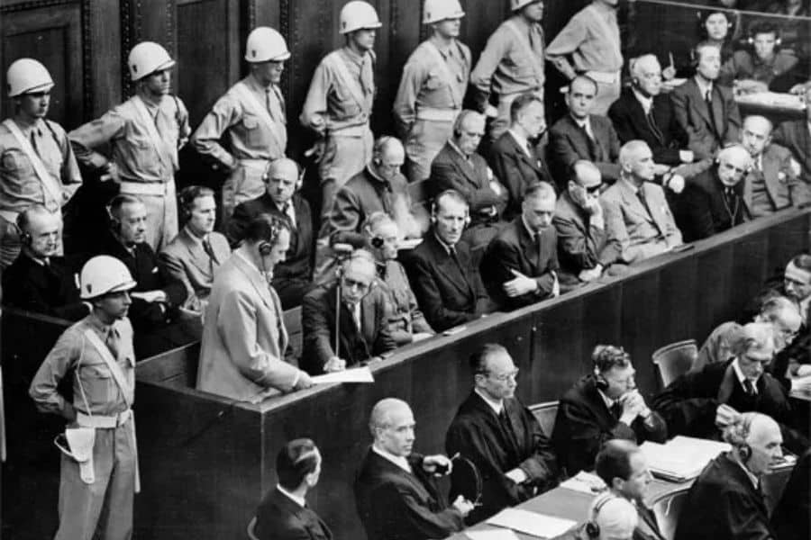 Courtroom scene from the Nuremberg Trials with defendants seated in a guarded dock while soldiers wearing helmets stand behind them. Lawyers, judges, and officials fill the courtroom during the war crimes proceedings in Nuremberg.