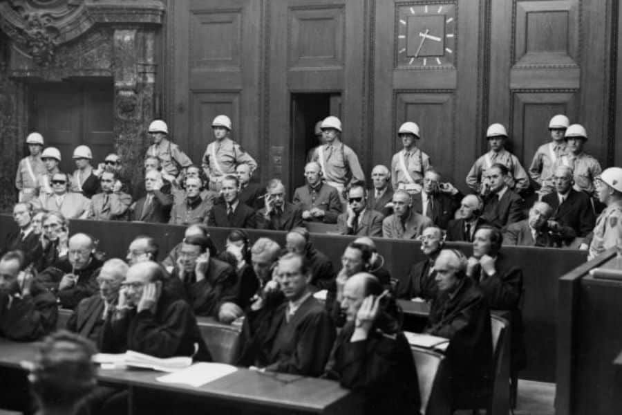 Wide view of the Nuremberg courtroom showing rows of judges, lawyers, translators, and guarded defendants during the postwar trials. Military guards line the walls while attendees wear headphones for multilingual translation during the Nuremberg proceedings.