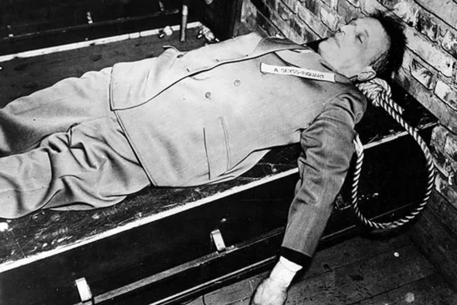 Black and white historical photograph showing a deceased man lying on a bench with a rope around his neck after execution following the Nuremberg Trials. The stark image documents the aftermath of sentences carried out against convicted Nazi officials in Nuremberg.