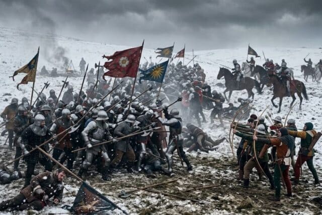 Large scale medieval battle taking place in a snowy field with armored infantry clashing, archers firing arrows, and mounted knights charging through smoke and chaos. Colorful banners wave above the fighting as soldiers engage in close combat during the intense winter battle.