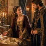 A noblewoman in a burgundy and gold gown gestures toward a sleeping baby in an ornate wooden cradle while a richly dressed nobleman looks on. The candlelit stone chamber and royal clothing evoke the world behind traditional medieval girls names.