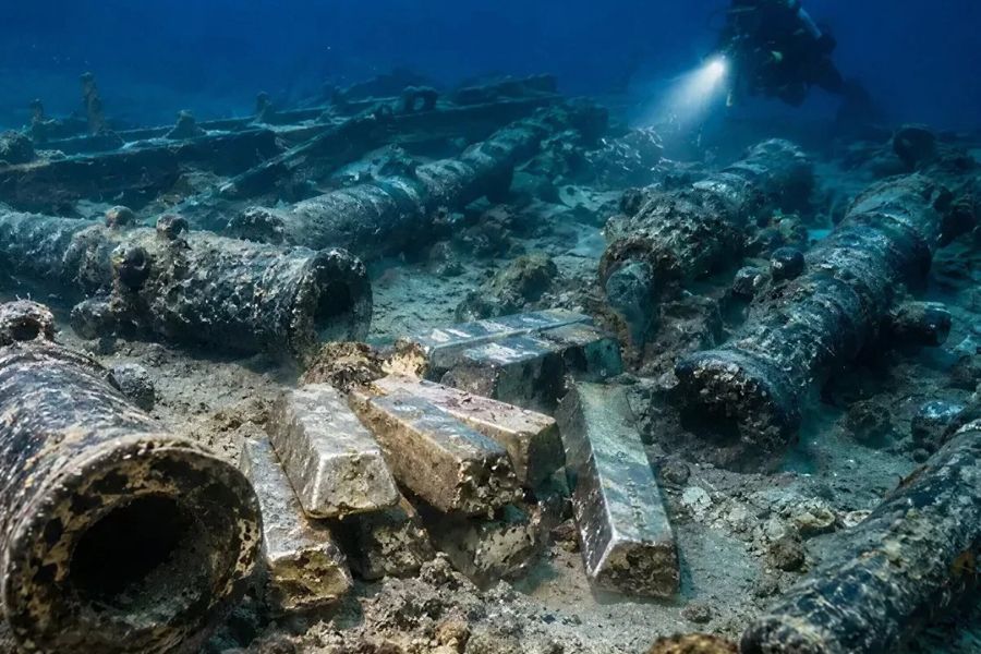 Corroded cannons and silver ingots rest on the seafloor beside a historic shipwreck while a diver shines a light across the site. The underwater discovery highlights maritime history connected to Cadiz and the valuable cargo carried by ships sailing its trade routes.