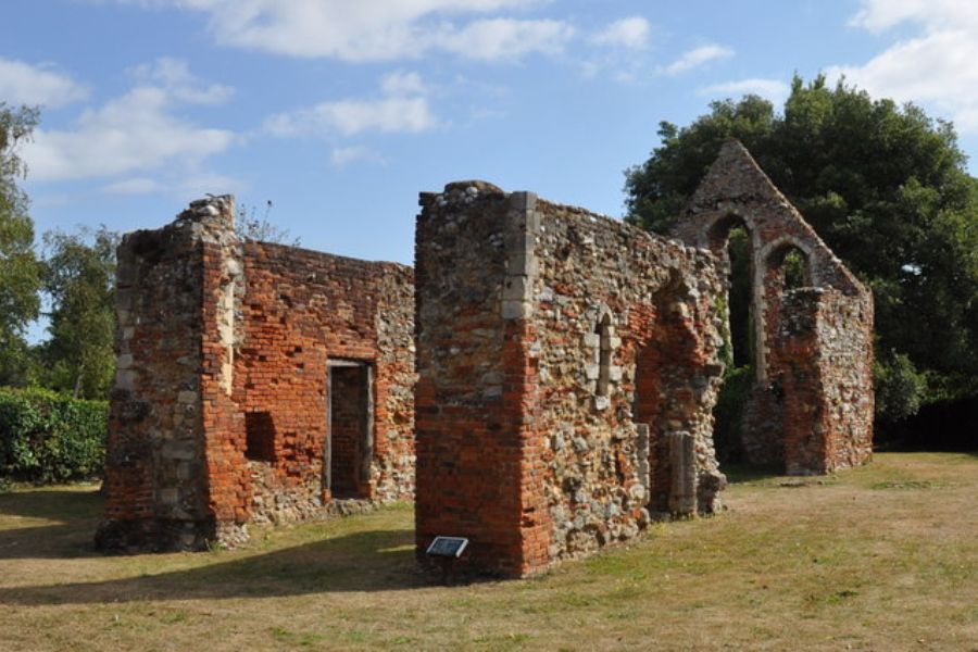 Roofless brick and flint ruins of a medieval hospital associated with the care and isolation of lepers. Arched window openings and broken walls remain on a grassy site beneath a blue sky.
