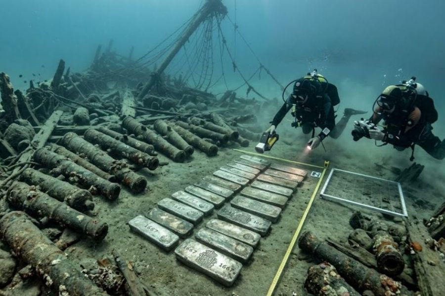Two scuba divers document rows of silver ingots and centuries old cannons scattered around a submerged shipwreck. The archaeological survey showcases a remarkable underwater treasure site linked to the seafaring history of Cadiz.