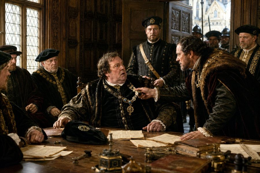 A group of men in Tudor times of Henry VIII sitting around a table and one man leaning over and reading towards Thomas Cromwell