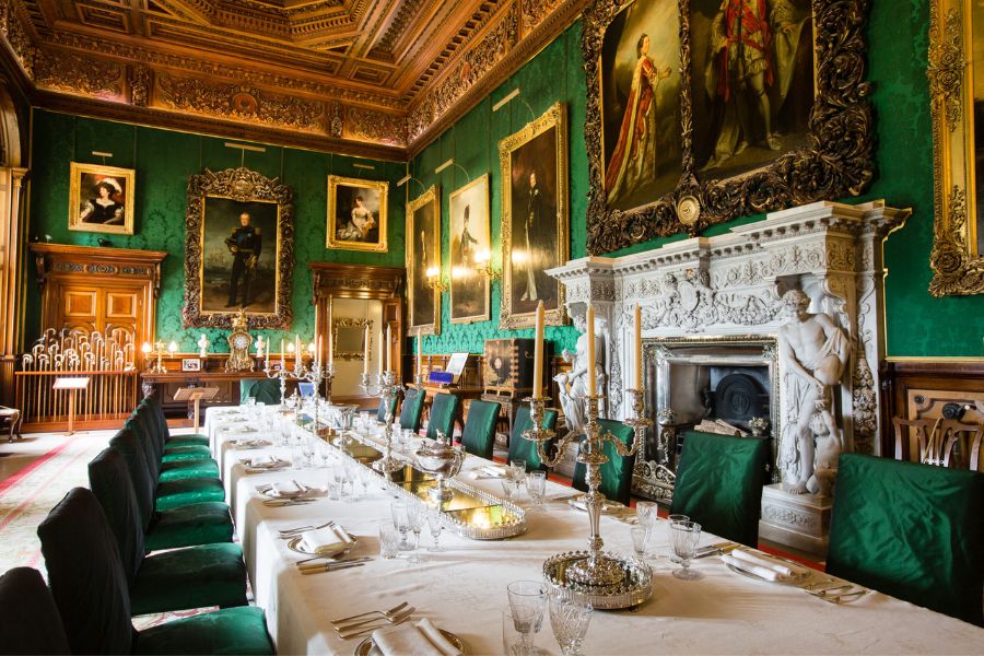 Long banquet table set with crystal glassware inside an ornate room with green walls gold frames and large portraits. This grand interior shows the stately side of English castles and historic royal rooms.